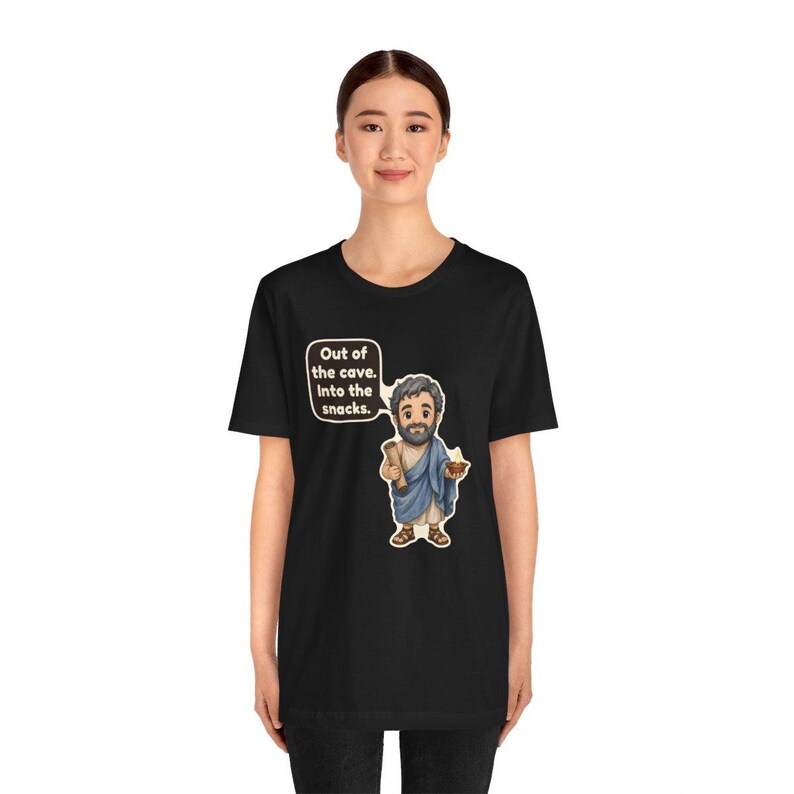 Peut inclure: Un t-shirt noir avec une illustration de dessin anim&eacute; repr&eacute;sentant un homme barbu en toge bleue tenant un parchemin et un bol. Une bulle au-dessus du personnage contient le texte : Out of the cave. Into the snacks. Le motif est centr&eacute;.
