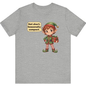 May include: A heather gray t-shirt featuring a cartoon elf wearing a green and red outfit with a matching hat. The elf holds a red gift box with a green ribbon. A speech bubble above the elf contains the text: Not short. Seasonally compact.