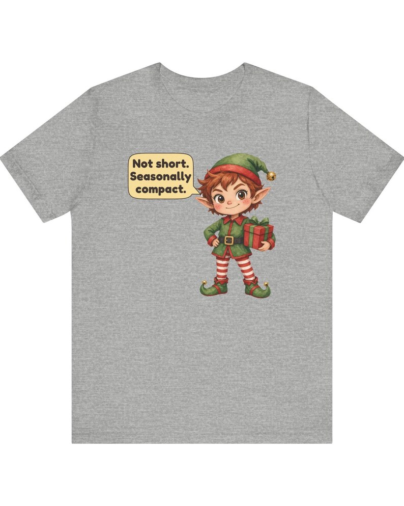 May include: A heather gray t-shirt featuring a cartoon elf wearing a green and red outfit with a matching hat. The elf holds a red gift box with a green ribbon. A speech bubble above the elf contains the text: Not short. Seasonally compact.