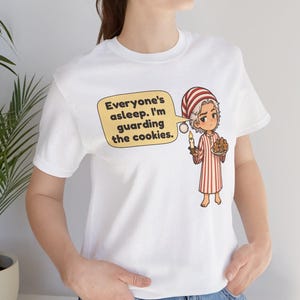 Op de afbeelding: Een wit T-shirt met korte mouwen en een cartoonillustratie van een persoon in een rood-wit gestreepte pyjama en een bijpassende slaapmuts. Het personage houdt een kaars en een bord met koekjes vast met een tekstballon, Everyone's asleep. I'm guarding the cookies.