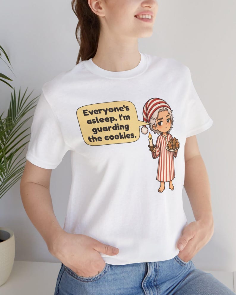 Op de afbeelding: Een wit T-shirt met korte mouwen en een cartoonillustratie van een persoon in een rood-wit gestreepte pyjama en een bijpassende slaapmuts. Het personage houdt een kaars en een bord met koekjes vast met een tekstballon, Everyone's asleep. I'm guarding the cookies.
