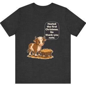 以下が含まれることがあります： ダークチャコールグレーのTシャツ。干し草が入った木製の飼い葉桶の横に立つ、茶色と白の牛の漫画イラストが描かれています。牛の上の吹き出しには「Hosted the first Christmas. No thank-you note.」というテキストがあります。