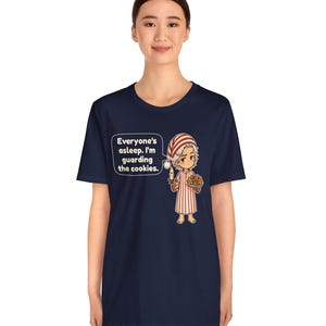Op de afbeelding: Een marineblauw t-shirt met een cartoonillustratie van een persoon in een rood-wit gestreepte pyjama die een kaars en een bord met koekjes vasthoudt. De tekst luidt: Everyone's asleep. I'm guarding the cookies.