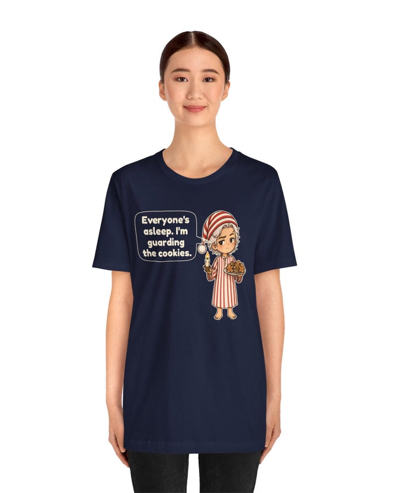 Op de afbeelding: Een marineblauw t-shirt met een cartoonillustratie van een persoon in een rood-wit gestreepte pyjama die een kaars en een bord met koekjes vasthoudt. De tekst luidt: Everyone's asleep. I'm guarding the cookies.