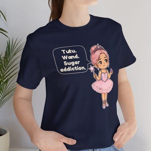May include: A navy blue t-shirt featuring a graphic of a cartoon ballerina in a pink tutu and crown holding a star wand. A speech bubble next to the character contains the text: Tutu. Wand. Sugar addiction. The design is centered on the chest.