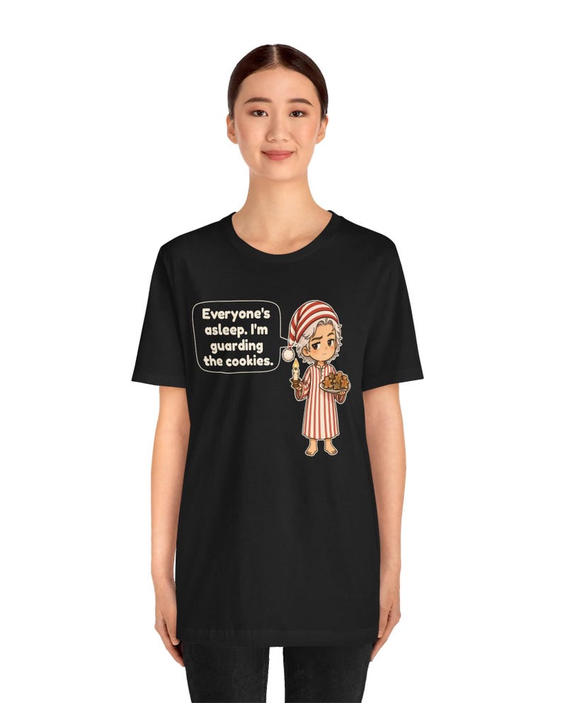 Op de afbeelding: Een zwart t-shirt met een cartoonillustratie van een figuur in een rood-wit gestreepte pyjama en bijpassende slaapmuts, met een kaars en een bord koekjes, met de tekst: Everyone's asleep. I'm guarding the cookies.