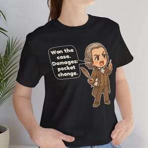 May include: A black t-shirt featuring a cartoon illustration of a historical figure in a brown suit holding a quill. A speech bubble contains the text: Won the case. Damages: pocket change. The figure has a powdered wig and a determined expression.