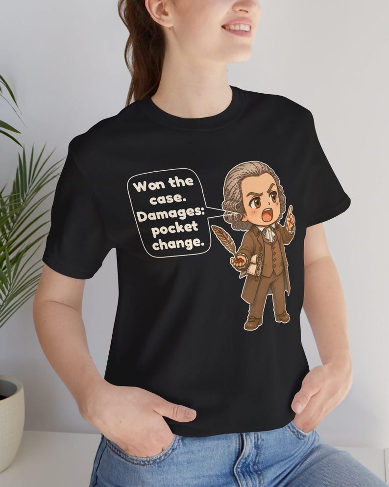 May include: A black t-shirt featuring a cartoon illustration of a historical figure in a brown suit holding a quill. A speech bubble contains the text: Won the case. Damages: pocket change. The figure has a powdered wig and a determined expression.