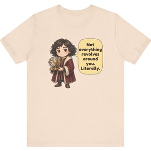 May include: A beige short-sleeve t-shirt featuring a cartoon character with dark curly hair wearing a maroon coat with white fur trim. The character holds a gold model of a solar system. A speech bubble contains the text: Not everything revolves around you. Literally.