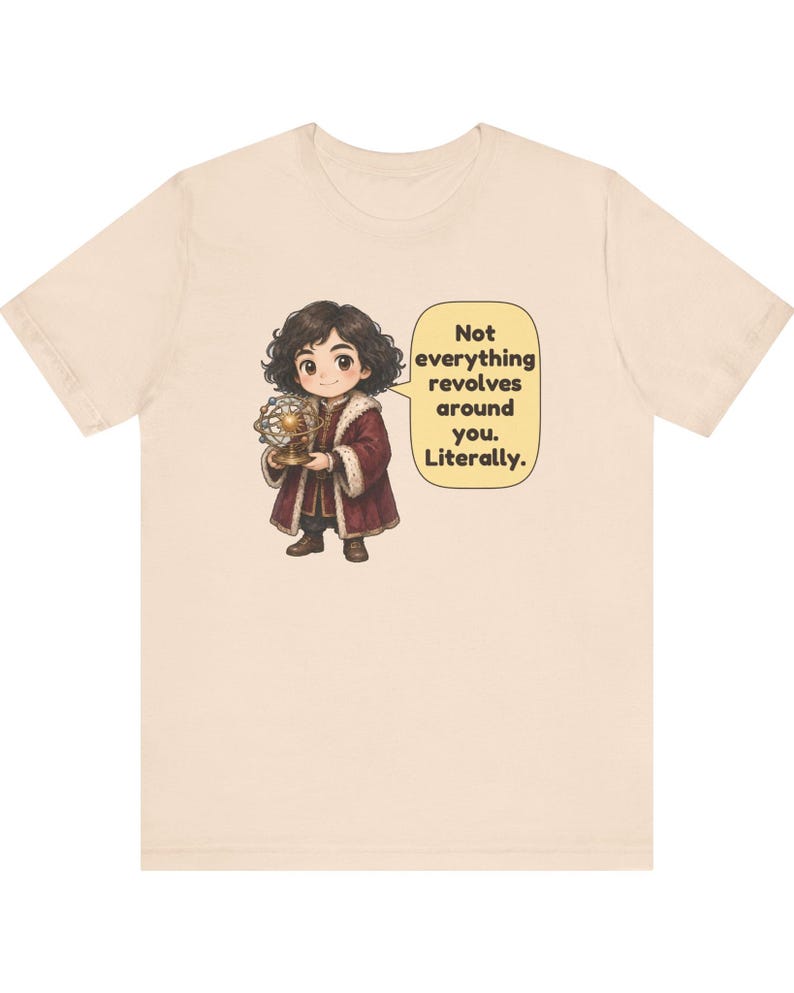 May include: A beige short-sleeve t-shirt featuring a cartoon character with dark curly hair wearing a maroon coat with white fur trim. The character holds a gold model of a solar system. A speech bubble contains the text: Not everything revolves around you. Literally.