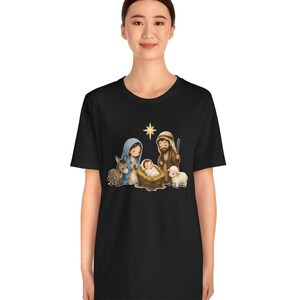 May include: A black short-sleeve t-shirt featuring a colorful cartoon-style graphic of a nativity scene. The print depicts a baby in a manger, a woman in a blue head covering, a man with a staff, a donkey, a sheep, and a yellow star.