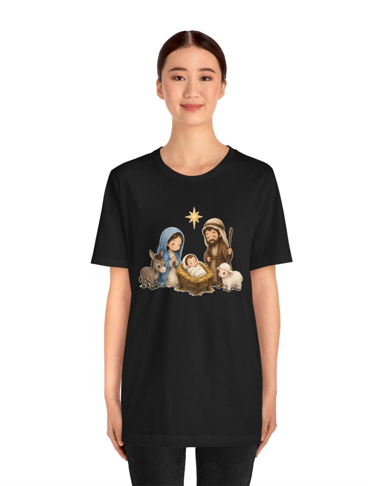 May include: A black short-sleeve t-shirt featuring a colorful cartoon-style graphic of a nativity scene. The print depicts a baby in a manger, a woman in a blue head covering, a man with a staff, a donkey, a sheep, and a yellow star.