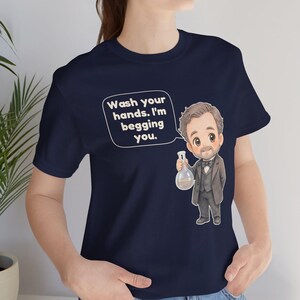 May include: A navy blue t-shirt featuring a cartoon illustration of a man in a suit holding a glass flask. A speech bubble above the figure contains the text: Wash your hands. I'm begging you. The design is centered on the front of the garment.