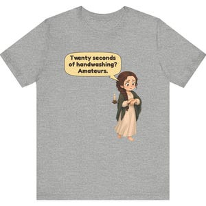 Op de afbeelding: Een grijs gem&ecirc;leerd t-shirt met een cartoonillustratie van een persoon met lang bruin haar in een vlecht, gekleed in een cr&egrave;mekleurige jurk en een donkere sjaal, met een brandende kaars. Een tekstballon bevat de tekst: Twenty seconds of handwashing? Amateurs.