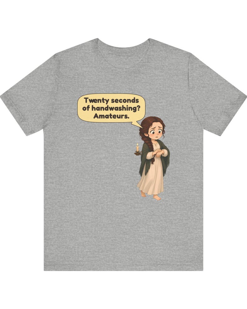 Op de afbeelding: Een grijs gem&ecirc;leerd t-shirt met een cartoonillustratie van een persoon met lang bruin haar in een vlecht, gekleed in een cr&egrave;mekleurige jurk en een donkere sjaal, met een brandende kaars. Een tekstballon bevat de tekst: Twenty seconds of handwashing? Amateurs.