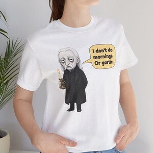 May include: A white t-shirt featuring a graphic illustration of a vampire with long gray hair, wearing a black coat, holding a lit candle, and a speech bubble that reads, "I don't do mornings. Or garlic." The shirt has a crew neck and short sleeves.
