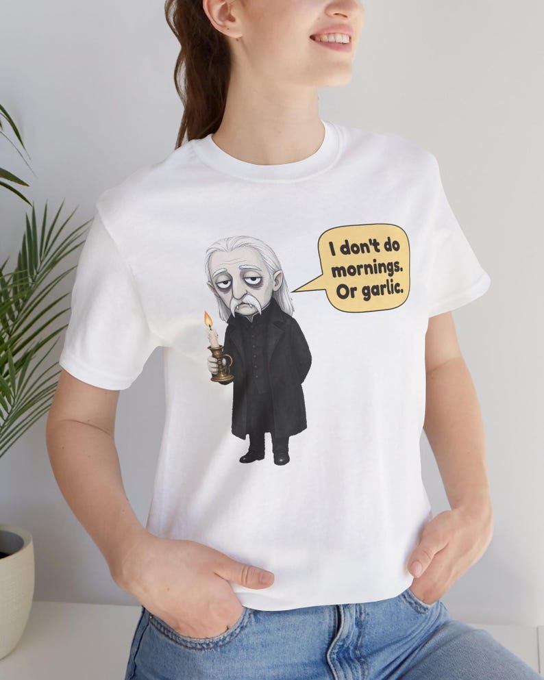 May include: A white t-shirt featuring a graphic illustration of a vampire with long gray hair, wearing a black coat, holding a lit candle, and a speech bubble that reads, "I don't do mornings. Or garlic." The shirt has a crew neck and short sleeves.