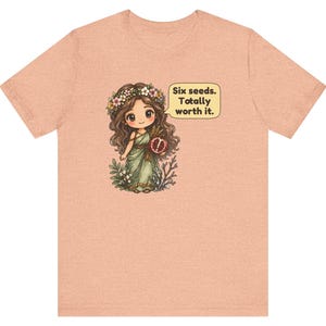 May include: A peach-colored t-shirt featuring a cartoon illustration of a character with long brown hair, a flower crown, and a green dress holding a pomegranate. A speech bubble next to the character contains the text: Six seeds. Totally worth it.