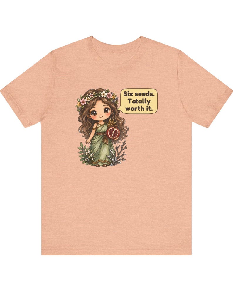May include: A peach-colored t-shirt featuring a cartoon illustration of a character with long brown hair, a flower crown, and a green dress holding a pomegranate. A speech bubble next to the character contains the text: Six seeds. Totally worth it.