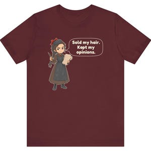 May include: A maroon short-sleeve t-shirt featuring a cartoon illustration of a character with a red hair bow, brown dress, and apron, holding a quill and paper. A speech bubble contains the text: Sold my hair. Kept my opinions.