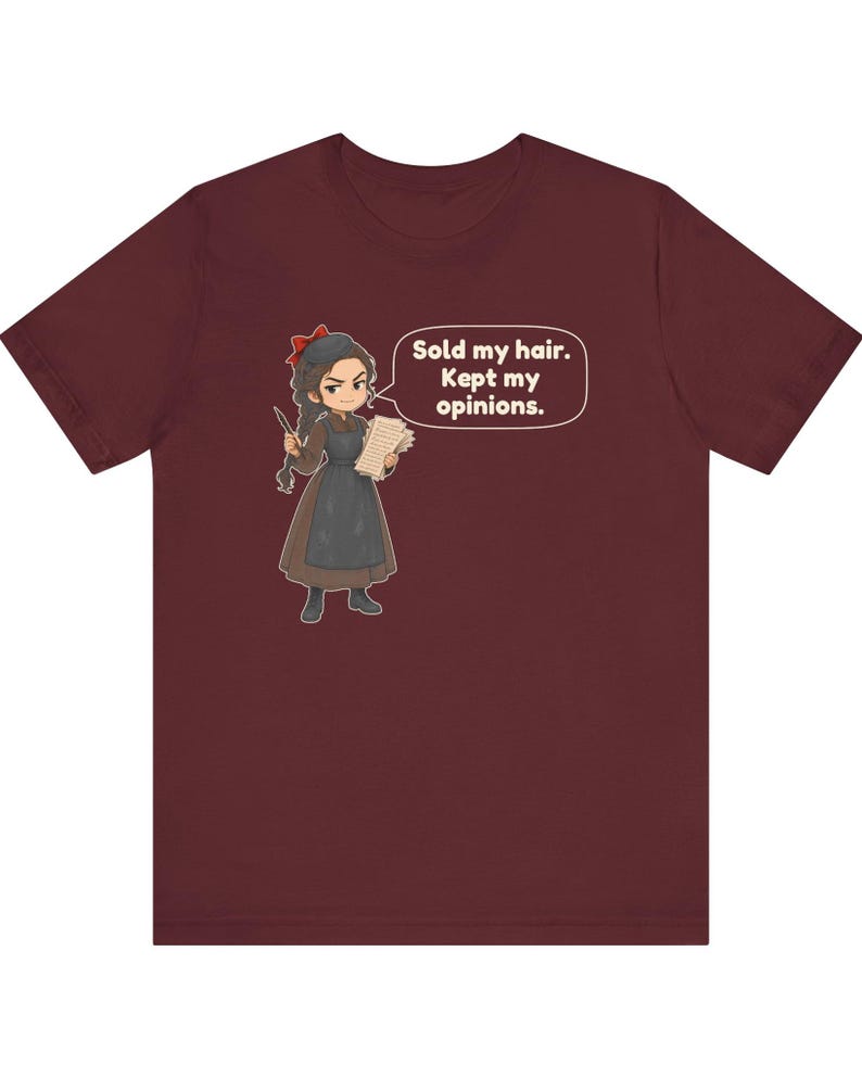 May include: A maroon short-sleeve t-shirt featuring a cartoon illustration of a character with a red hair bow, brown dress, and apron, holding a quill and paper. A speech bubble contains the text: Sold my hair. Kept my opinions.