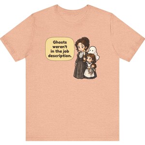 Op de afbeelding: Een perzikkleurig T-shirt met korte mouwen en een afbeelding van een persoon in een donkere jurk met een kaars, een kind met een pop en een klein wit spookje. Een tekstballon bevat de tekst: Ghosts weren't in the job description.