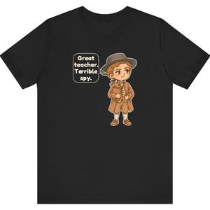 May include: A black t-shirt featuring a cartoon illustration of a person in 18th-century attire, including a brown coat, vest, and hat. A speech bubble next to the character contains the text: Great teacher. Terrible spy.