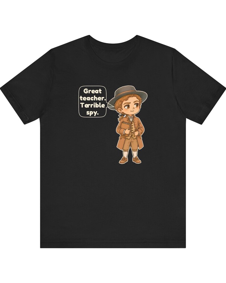May include: A black t-shirt featuring a cartoon illustration of a person in 18th-century attire, including a brown coat, vest, and hat. A speech bubble next to the character contains the text: Great teacher. Terrible spy.
