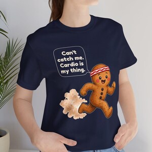 May include: A navy blue t-shirt featuring a graphic of a running gingerbread person wearing a red and white headband. A speech bubble above the character contains the text: Can't catch me. Cardio is my thing. A cloud of dust trails behind the figure.