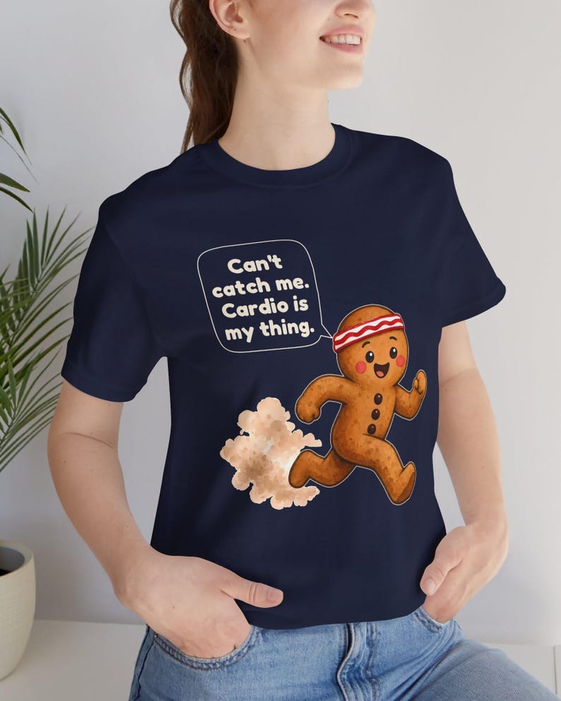 May include: A navy blue t-shirt featuring a graphic of a running gingerbread person wearing a red and white headband. A speech bubble above the character contains the text: Can't catch me. Cardio is my thing. A cloud of dust trails behind the figure.