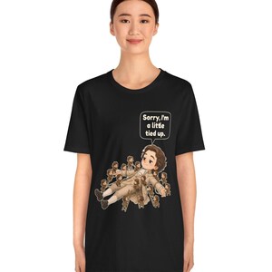 Op de afbeelding: Een zwart t-shirt met korte mouwen en een afbeelding van een liggend personage dat wordt vastgebonden door vele kleine figuurtjes. Boven het personage staat een witte tekstballon met de tekst, Sorry, I'm a little tied up.