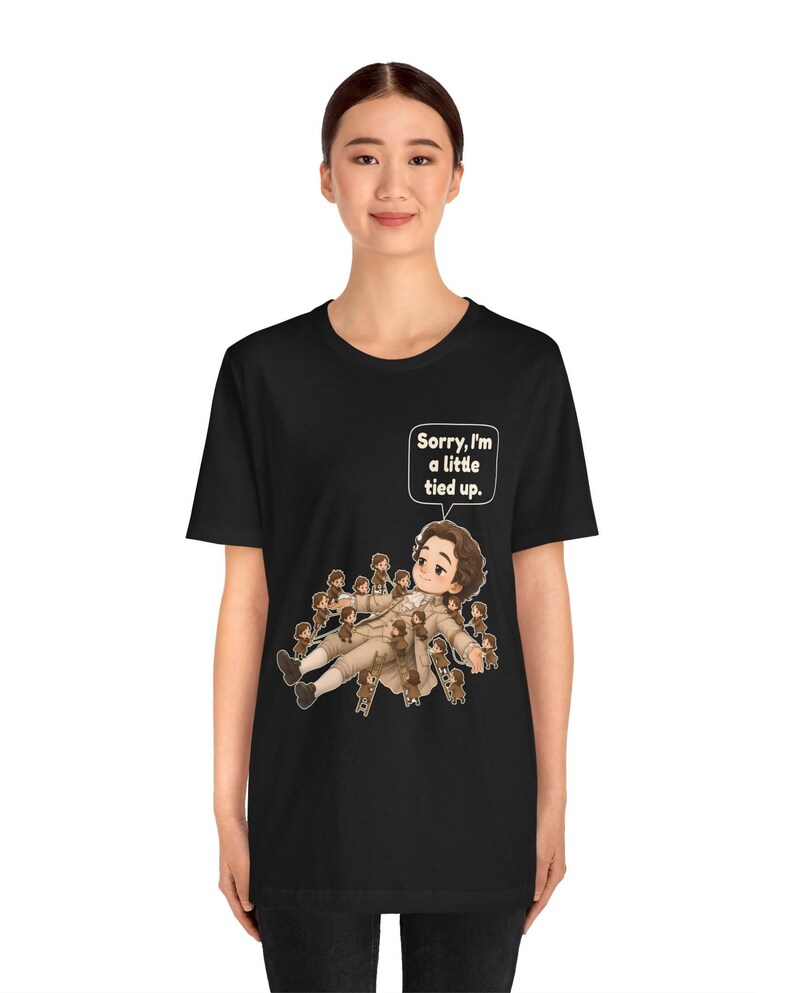 Op de afbeelding: Een zwart t-shirt met korte mouwen en een afbeelding van een liggend personage dat wordt vastgebonden door vele kleine figuurtjes. Boven het personage staat een witte tekstballon met de tekst, Sorry, I'm a little tied up.