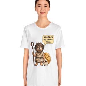 May include: A white t-shirt featuring a cartoon cyclops holding a wooden staff and standing next to a small sheep and a large wheel of cheese. A speech bubble above the cyclops contains the text: Guests ate my cheese. Rude.