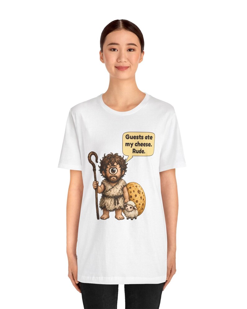 May include: A white t-shirt featuring a cartoon cyclops holding a wooden staff and standing next to a small sheep and a large wheel of cheese. A speech bubble above the cyclops contains the text: Guests ate my cheese. Rude.