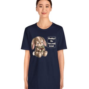 May include: A navy blue short-sleeve t-shirt featuring a graphic of a brown stuffed bunny holding a sprig of holly. Next to the bunny is a speech bubble with the text: Shabby? No. Very well loved. The design is centered on the chest.