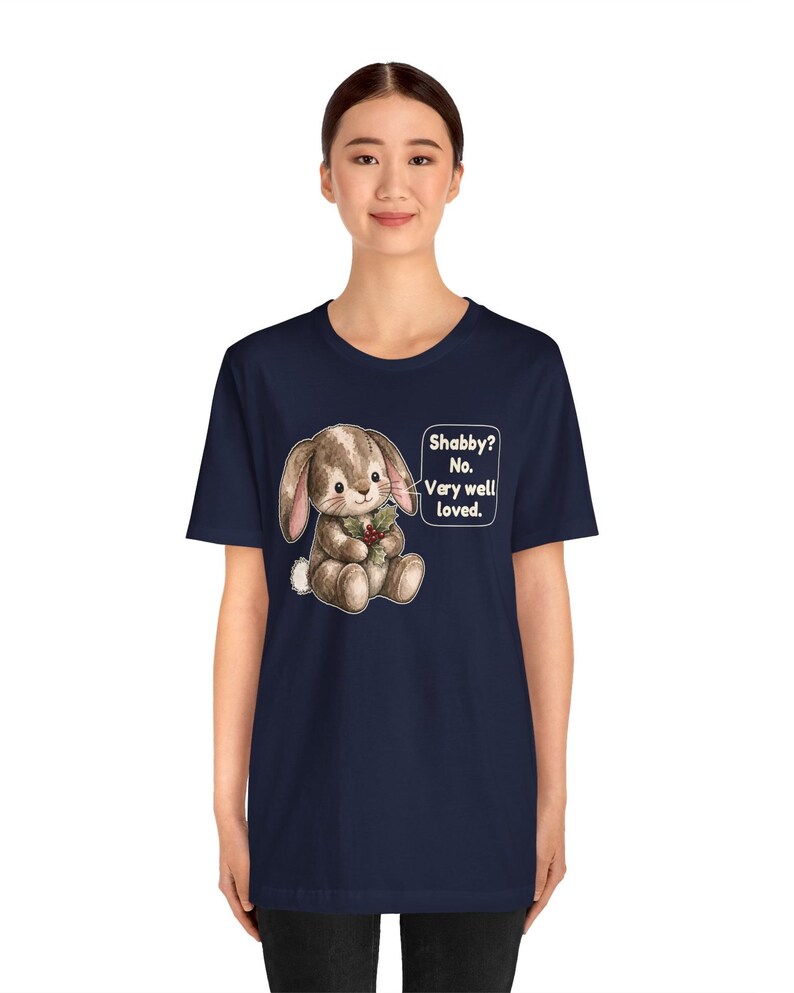 May include: A navy blue short-sleeve t-shirt featuring a graphic of a brown stuffed bunny holding a sprig of holly. Next to the bunny is a speech bubble with the text: Shabby? No. Very well loved. The design is centered on the chest.