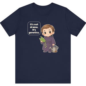 May include: A navy blue t-shirt featuring a cartoon illustration of a monk holding a pea pod and a watering can. A speech bubble next to the character contains the text: It's not drama. It's genetics. The design is centered on the chest.