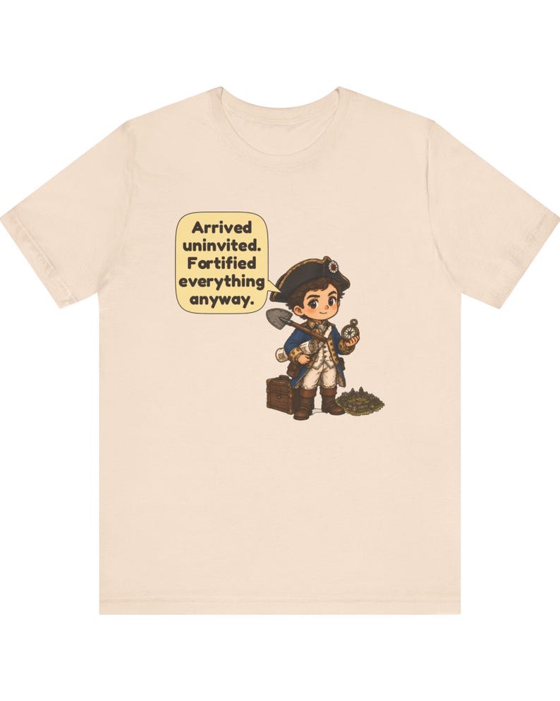 May include: A light beige t-shirt featuring a cartoon illustration of a historical explorer in a tricorn hat holding a shovel and compass. Next to the character is a speech bubble with the text: Arrived uninvited. Fortified everything anyway.