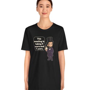 May include: A black t-shirt featuring a cartoon illustration of Abraham Lincoln wearing a top hat and holding a pocket watch. A speech bubble next to the figure contains the text: This meeting is taking 4 score and 7 years.