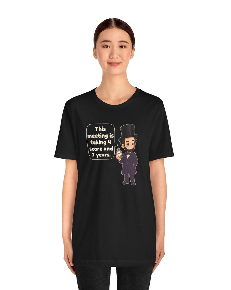 May include: A black t-shirt featuring a cartoon illustration of Abraham Lincoln wearing a top hat and holding a pocket watch. A speech bubble next to the figure contains the text: This meeting is taking 4 score and 7 years.