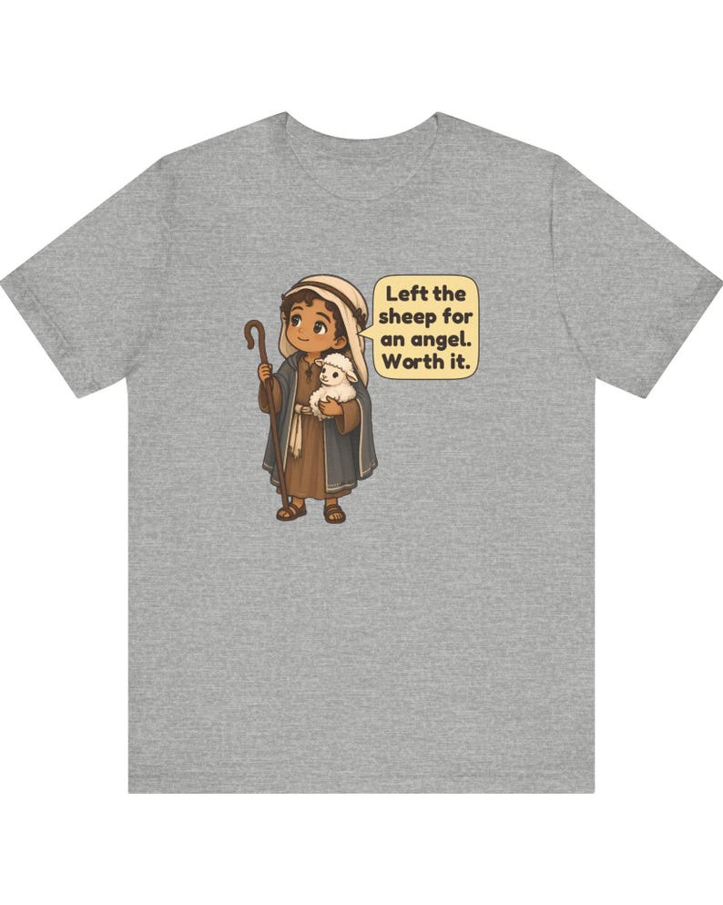 May include: A heather gray t-shirt featuring a cartoon illustration of a shepherd holding a small white sheep and a wooden staff. A speech bubble next to the figure contains the text: Left the sheep for an angel. Worth it.