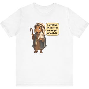 May include: A white t-shirt featuring a cartoon illustration of a shepherd holding a small white lamb and a wooden staff. A speech bubble next to the character contains the text: Left the sheep for an angel. Worth it.