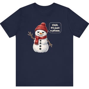 Pode incluir: Uma t-shirt azul-marinho de manga curta com o gr&aacute;fico de um boneco de neve a usar um gorro e um cachecol de malha vermelha. O boneco tem um nariz de cenoura, bra&ccedil;os de ramo e um bal&atilde;o com o texto: Chill. It's just a phase.
