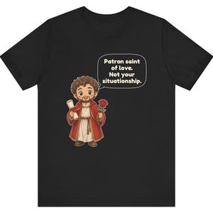 Puede incluir: Una camiseta negra con una ilustraci&oacute;n de dibujos animados de una figura barbuda con t&uacute;nica roja que sostiene un pergamino y una rosa roja. Un globo de texto sobre la figura contiene el texto: Patron saint of love. Not your situationship.