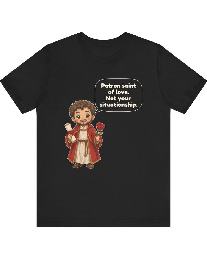 Puede incluir: Una camiseta negra con una ilustraci&oacute;n de dibujos animados de una figura barbuda con t&uacute;nica roja que sostiene un pergamino y una rosa roja. Un globo de texto sobre la figura contiene el texto: Patron saint of love. Not your situationship.