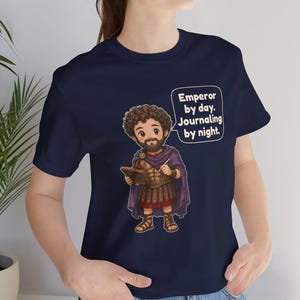 K&ouml;nnte beinhalten: Ein marineblaues T-Shirt mit einer Cartoon-Illustration eines b&auml;rtigen Kaisers in r&ouml;mischer R&uuml;stung, der ein Tagebuch und einen Stift h&auml;lt. Eine Sprechblase neben der Figur enth&auml;lt den Text: Emperor by day. Journaling by night.