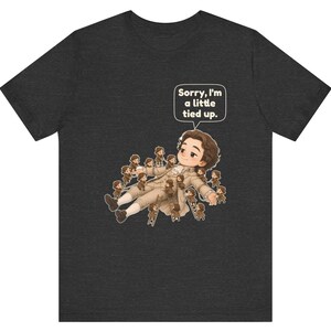Op de afbeelding: Een antracietgrijs t-shirt met een afbeelding van een groot persoon die ligt en omringd is door vele kleine figuurtjes die met ladders op hen klimmen. Een tekstballon bevat de tekst, Sorry, I'm a little tied up.