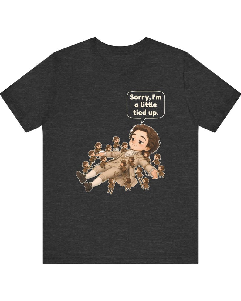 Op de afbeelding: Een antracietgrijs t-shirt met een afbeelding van een groot persoon die ligt en omringd is door vele kleine figuurtjes die met ladders op hen klimmen. Een tekstballon bevat de tekst, Sorry, I'm a little tied up.