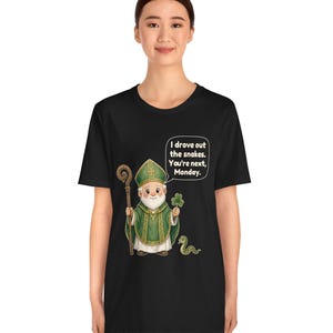 Puede incluir: Una camiseta negra con una ilustraci&oacute;n de dibujos animados de una figura con vestiduras religiosas verdes que sostiene un bast&oacute;n de madera y un tr&eacute;bol. Una peque&ntilde;a serpiente est&aacute; cerca. Un globo de texto contiene: I drove out the snakes. You're next, Monday.