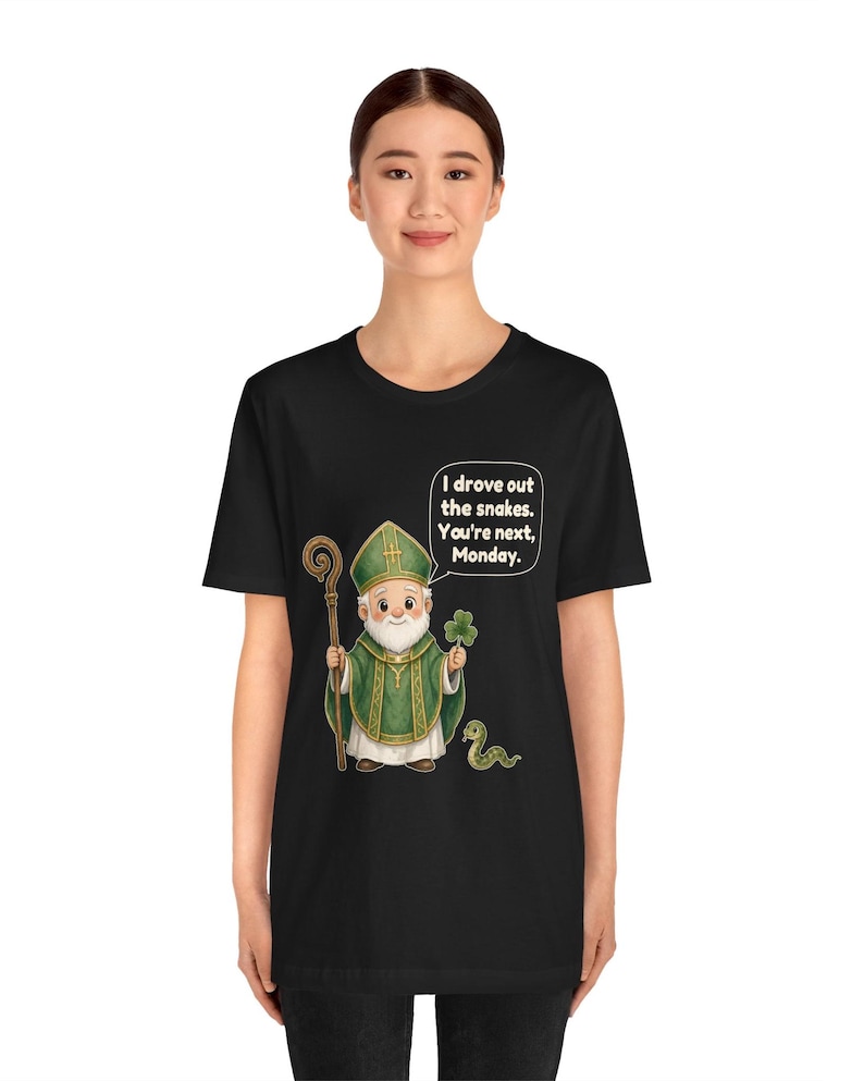 Puede incluir: Una camiseta negra con una ilustraci&oacute;n de dibujos animados de una figura con vestiduras religiosas verdes que sostiene un bast&oacute;n de madera y un tr&eacute;bol. Una peque&ntilde;a serpiente est&aacute; cerca. Un globo de texto contiene: I drove out the snakes. You're next, Monday.